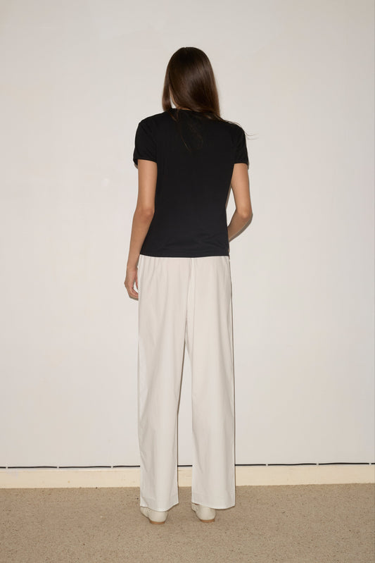A woman with straight brown hair faces an off-white wall, wearing the 0 Simple Tee - Black and relaxed white pants. The floor beneath her is carpeted in beige.