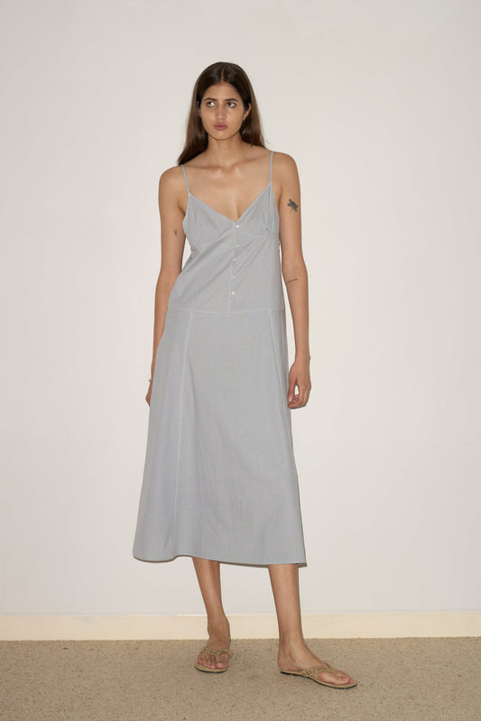 A woman with long brown hair wears the Double Seam Dress, a light blue, sleeveless, button-front midi dress with thin straps and beige sandals, standing against a plain light wall and beige floor.