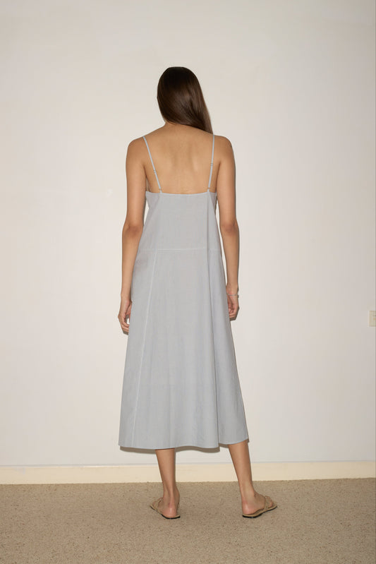 A woman with long brown hair stands facing an off-white wall, wearing a Deiji Studios Double Seam Dress - Sky Stripe with thin straps and beige sandals. Her arms are relaxed by her sides in the minimalist setting.