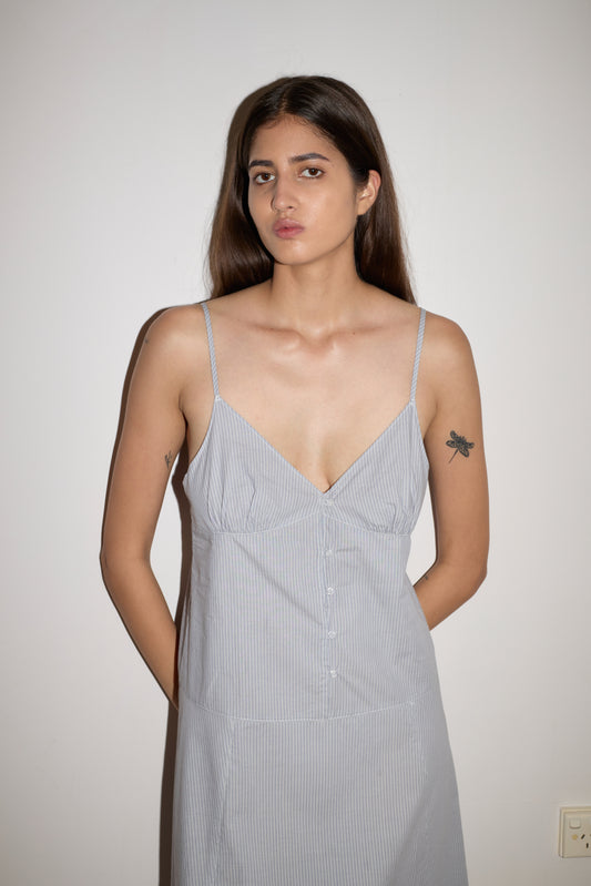 A woman with long brown hair stands against a white wall, wearing a Deiji Studios Double Seam Dress - Sky Stripe with thin straps. She has a dragonfly tattoo on her upper left arm and a neutral expression.