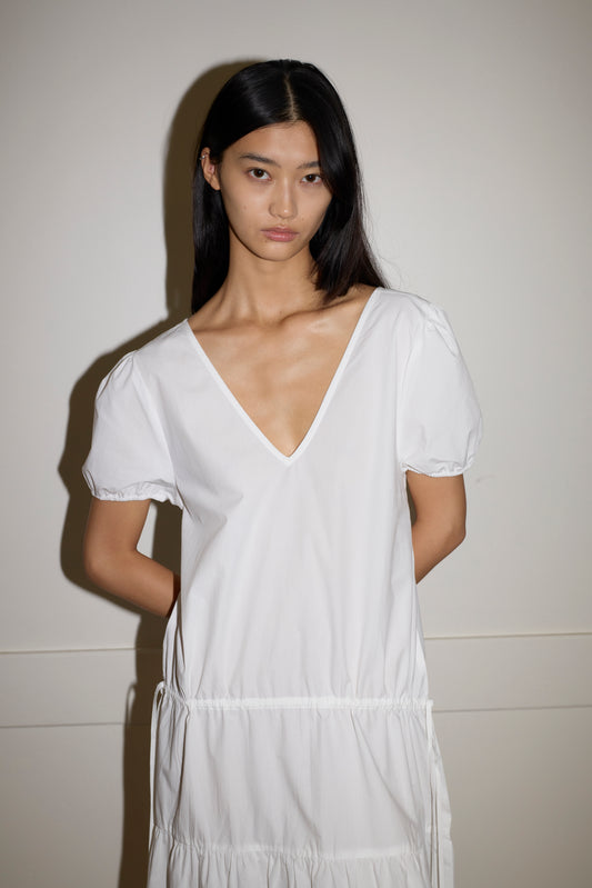 A model with long dark hair wears the Waistcord Dress, a white cotton piece with a deep V-neckline, short puff sleeves, and a gathered elastic waist, posing against a neutral wall.