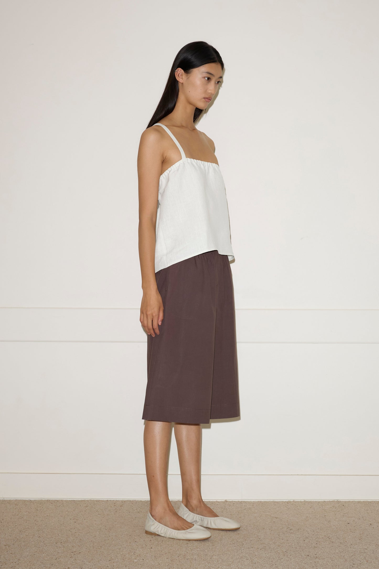 A woman with long dark hair stands sideways against a plain wall, wearing the Button Strap Top - White by Deiji Studios, loose brown knee-length culottes, and light-colored flat shoes.