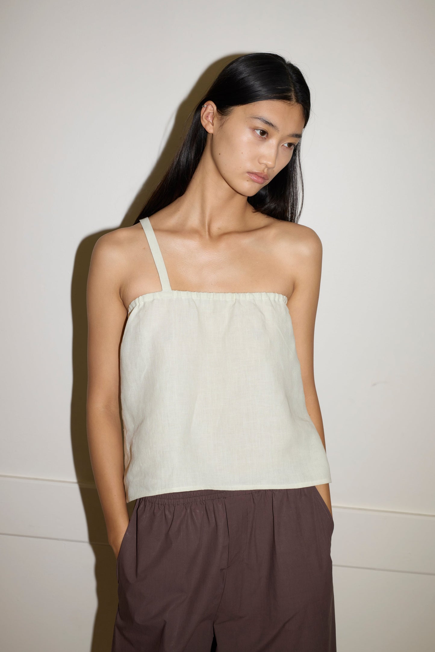 A woman with long, straight black hair wears the Button Strap Top in light beige and loose brown pants, standing against an off-white wall with her hands in her pockets, looking slightly downward.