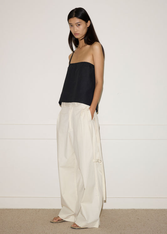 A woman with long dark hair stands against a white wall, gazing to the side. She wears a loose black strapless top, Deiji Studios Tab Pant in Off White, and beige sandals, with her hands casually placed in her pockets.