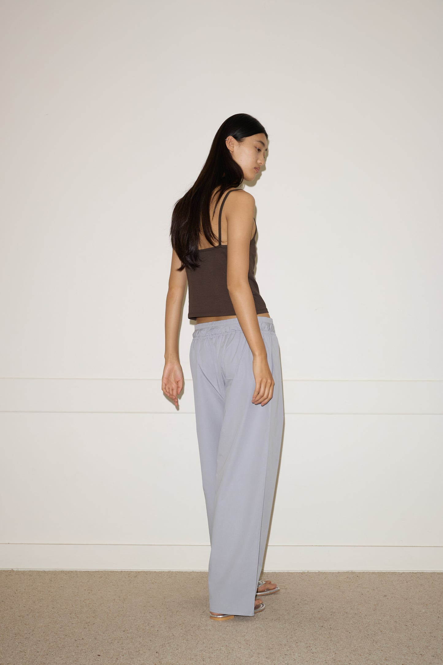 A woman with long dark hair, wearing the 0 Simple Tank - Caribou and light gray loose pants, stands in a minimalist room with a white wall and beige carpet, looking over her shoulder.