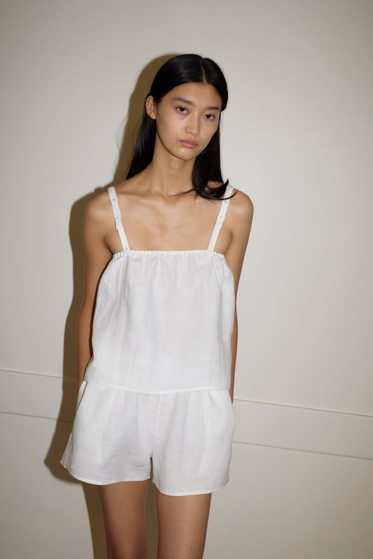 A woman with long dark hair wears the Button Linen Set— a sleeveless white top and matching shorts—while standing against a plain light background with a neutral expression.