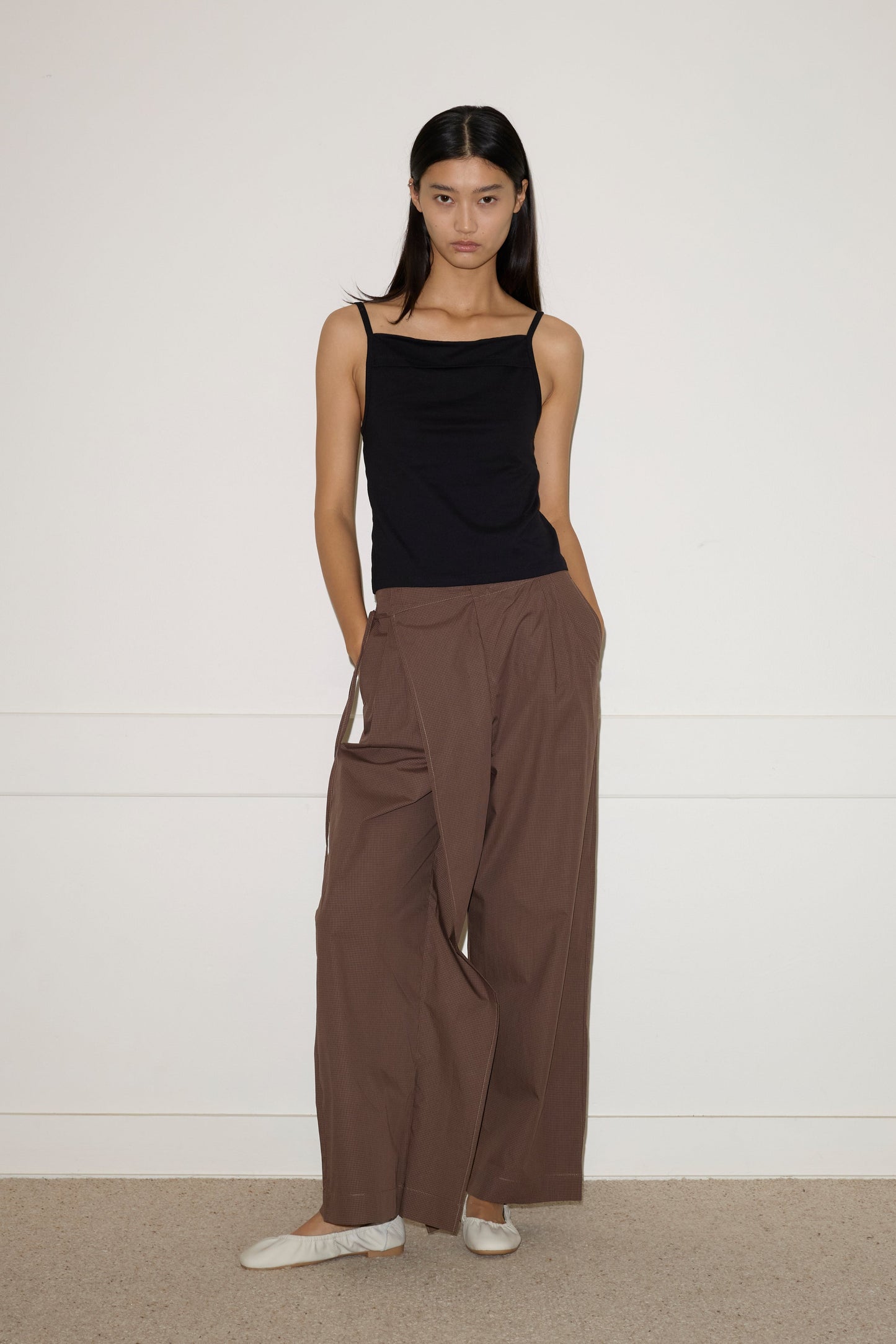 A woman stands against a white wall, wearing a black sleeveless top and Deiji Studios Double Layer Pant - Carob Check in organic cotton. She has long straight hair, white flats, and her hands are in her pockets.