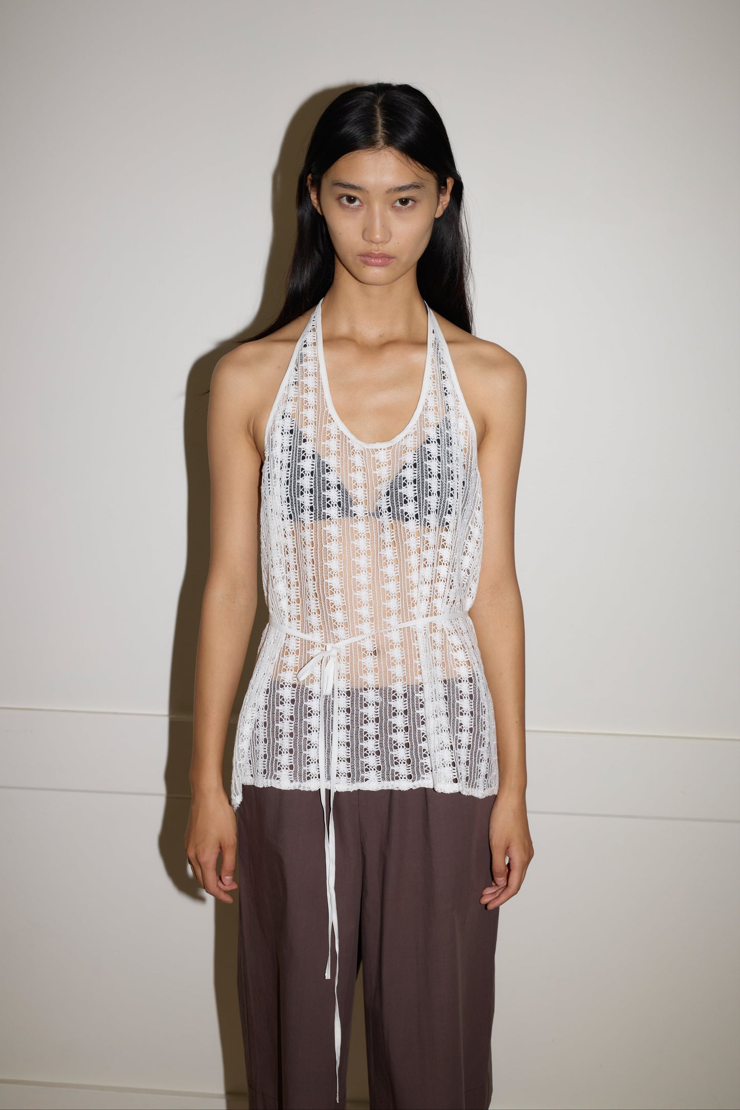 A person with long dark hair stands against a plain wall, wearing the 0 Thread Wrap Top in Off White over a black bralette and loose brown pants, looking directly at the camera with a neutral expression.