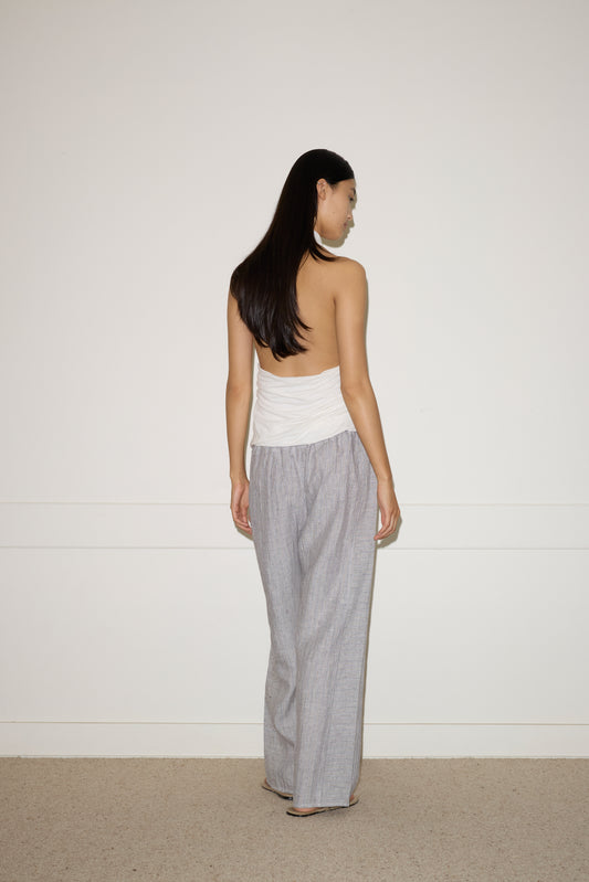 A woman with long dark hair faces away from the camera, wearing a white Jersey Top and loose gray pants against a plain white wall and beige carpeted floor.