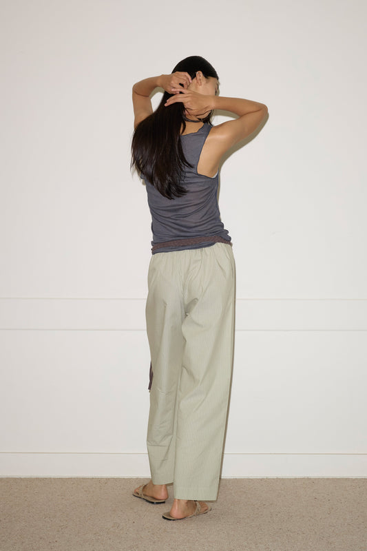 A person with long dark hair wears the Knit Tank - Steel by 0 and loose light pants, standing before a white wall with arms raised—an easy, effortless style in eco-friendly fashion.