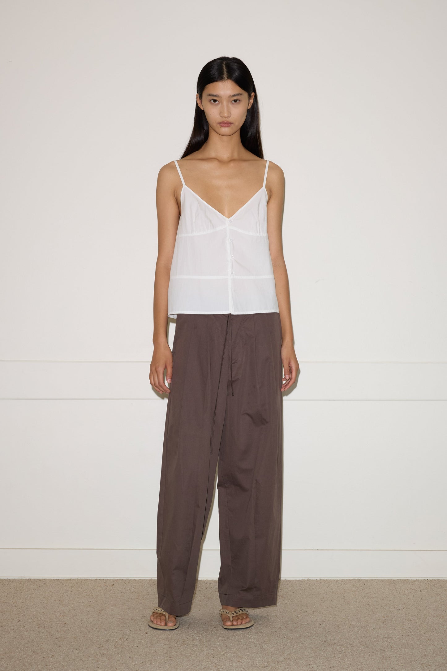 A woman with long straight hair and a neutral expression stands against a plain light wall, wearing the Seam Top - White by 0, loose brown pants, and sandals.