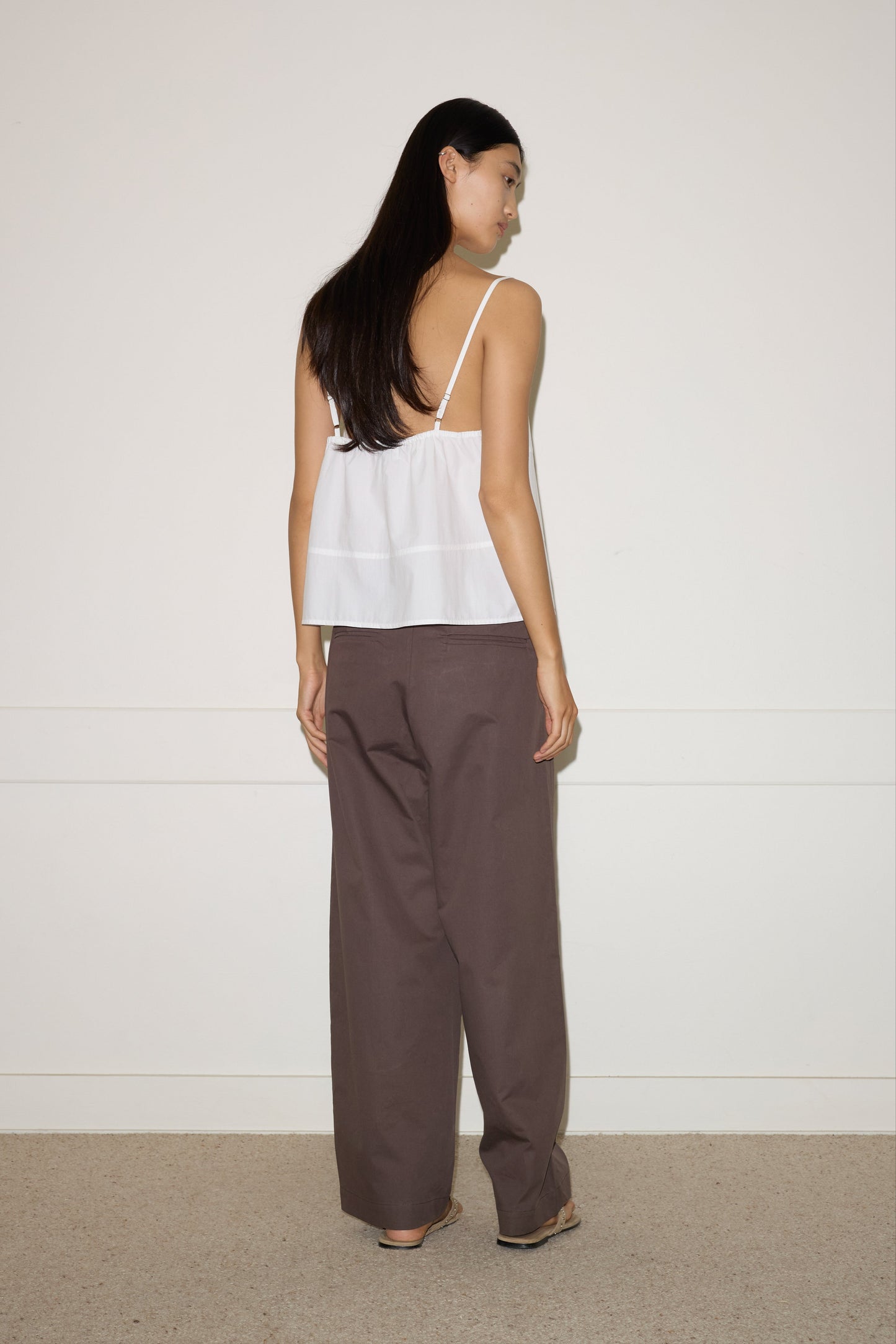 A woman with long dark hair, facing away, wears the Seam Top - White by 0 and loose brown pants against a plain white wall and beige carpet.