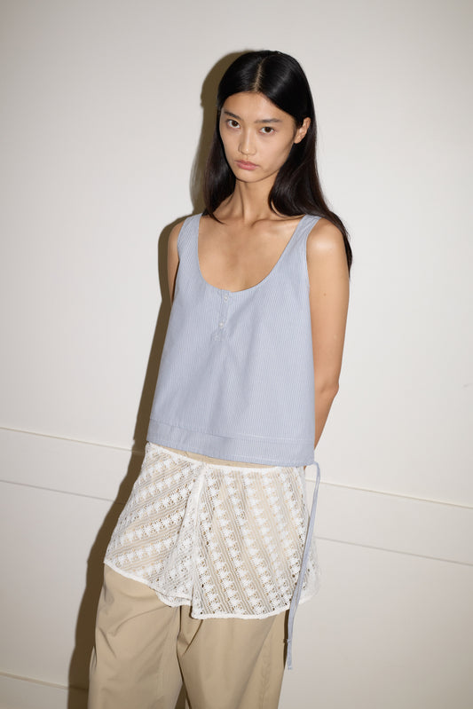 A person with long dark hair wears the Layered Top—a sleeveless light blue piece over a white lace layer—paired with beige pants, standing against a plain white wall with a neutral expression.