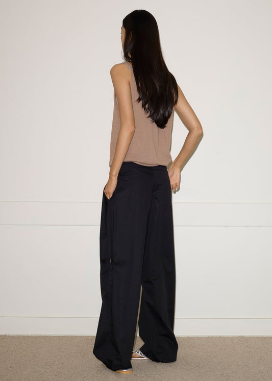 A person with long dark hair, wearing a sleeveless brown top and the Tab Pant - Black by Deiji Studios, stands indoors facing away from the camera against a plain light wall and carpeted floor.