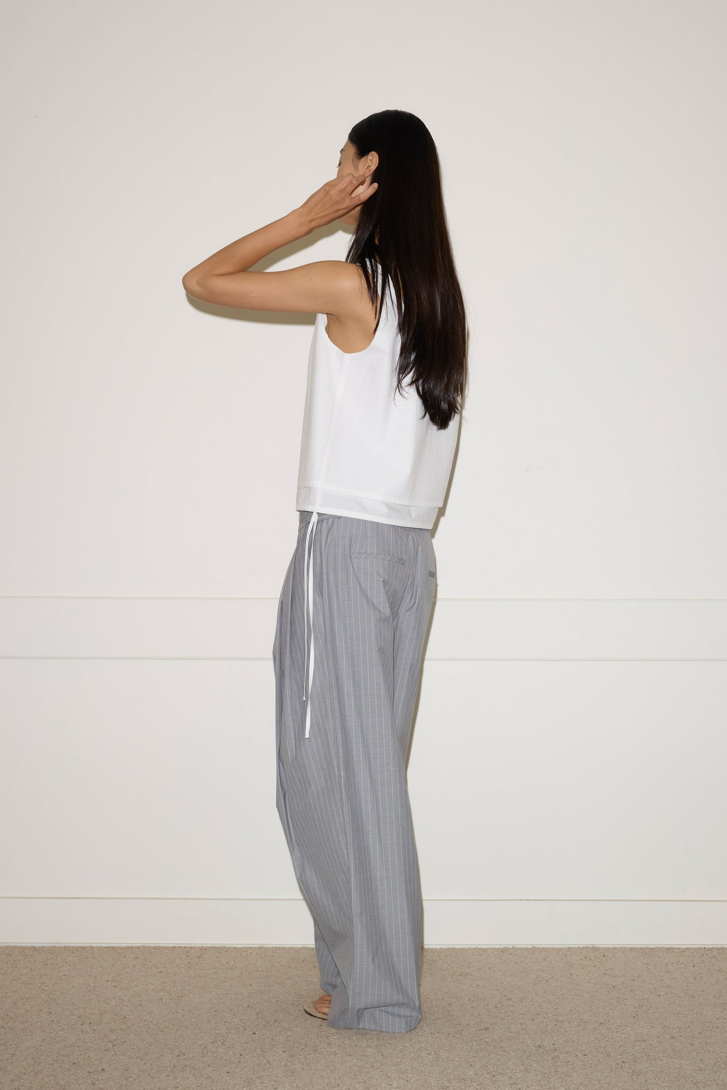 A person with long dark hair stands barefoot indoors, facing a white wall, wearing the 0 Layered Top - White and loose gray striped organic cotton pants. One hand is raised to their head.