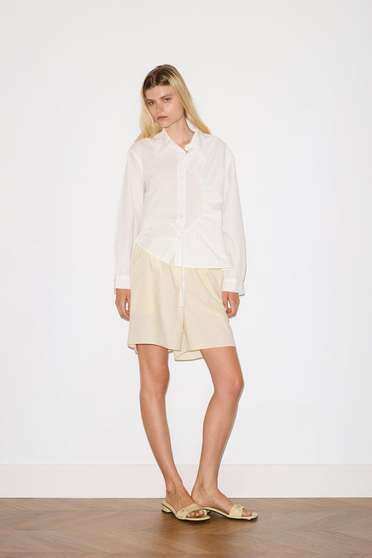 A woman stands against a plain white wall wearing the Arch Tie Shirt - White by Deiji Studios, paired with beige pinstripe knee-length shorts and beige sandals, her hair down and expression neutral.
