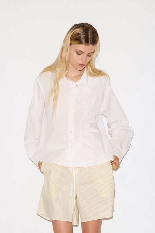 A woman with long blonde hair looks down while wearing the Arch Tie Shirt - White by Deiji Studios, paired with light pinstriped shorts. Her hands in her pockets, she stands against a plain white background.