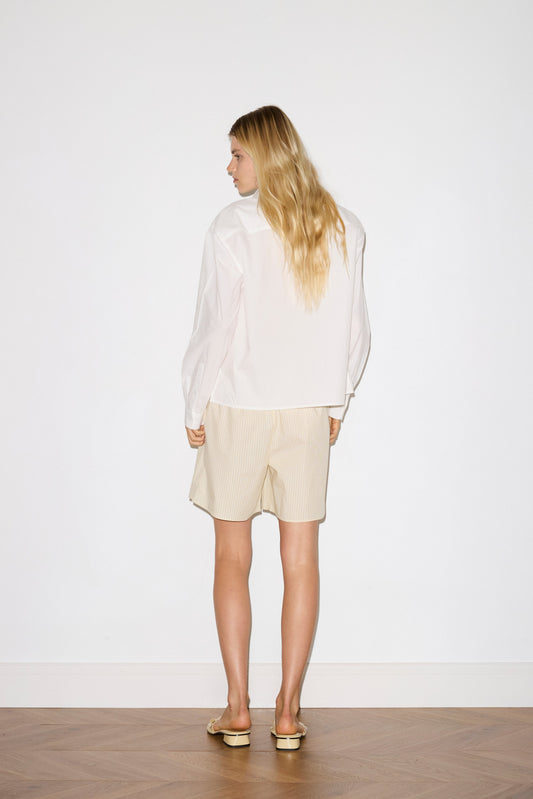 A person with long blonde hair stands facing away, wearing the Deiji Studios Arch Tie Shirt in white, paired with light beige shorts and sandals, in a minimal room with a white wall and wooden floor.