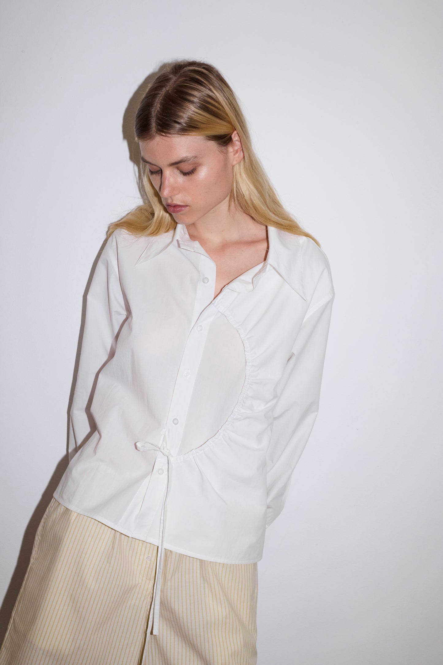 A woman with long blond hair looks down while wearing the Arch Tie Shirt - White by Deiji Studios, featuring an asymmetrical circular cutout, paired with beige striped pants against a plain white background.