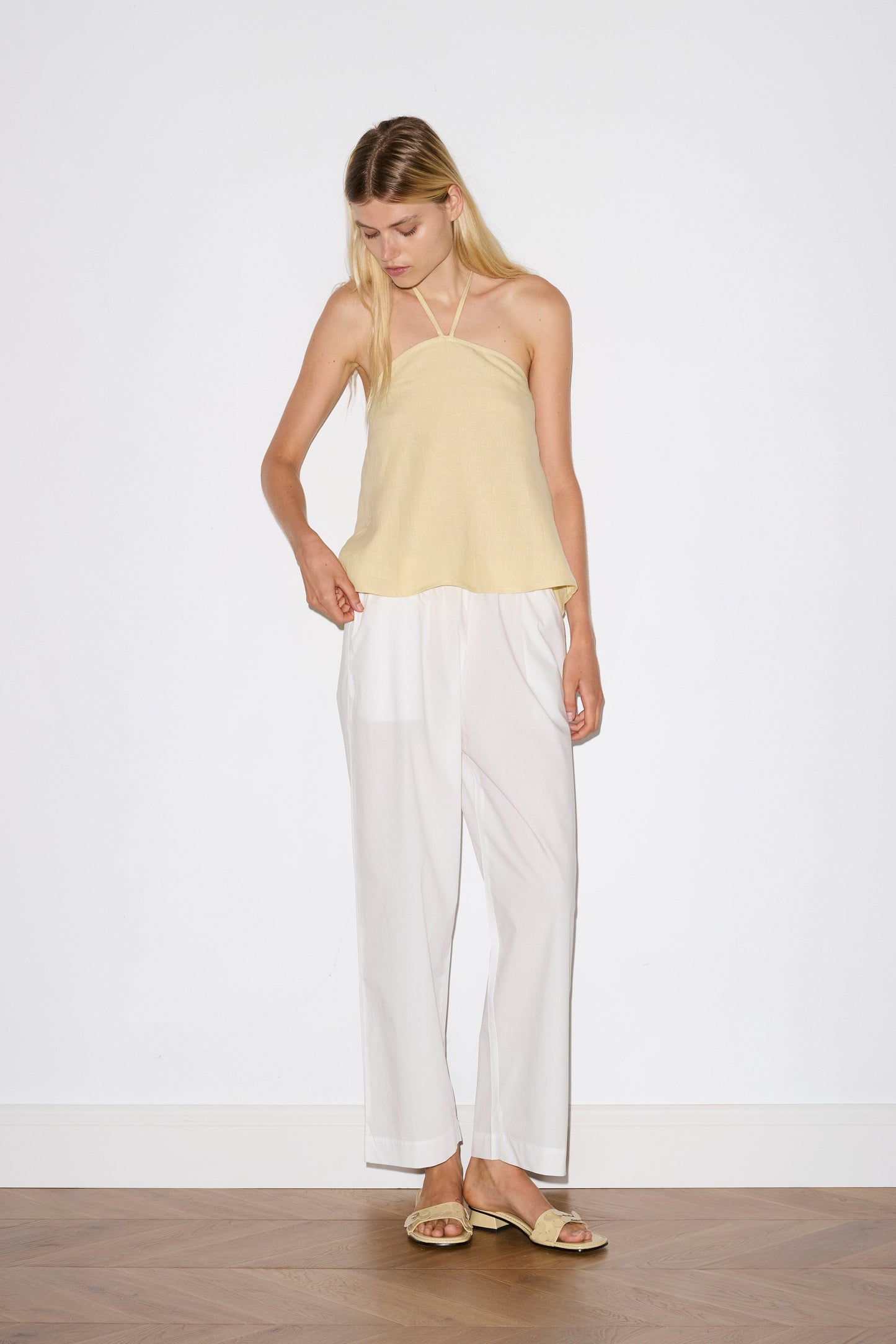 blonde haired female model wears a yellow relaxed halter top features flattering straight neckline, fine tie halter straps and snap closure at back with button detail, worn with a white ankle length pant features a soft minimal elastic waist with side seam pockets in a straight leg.