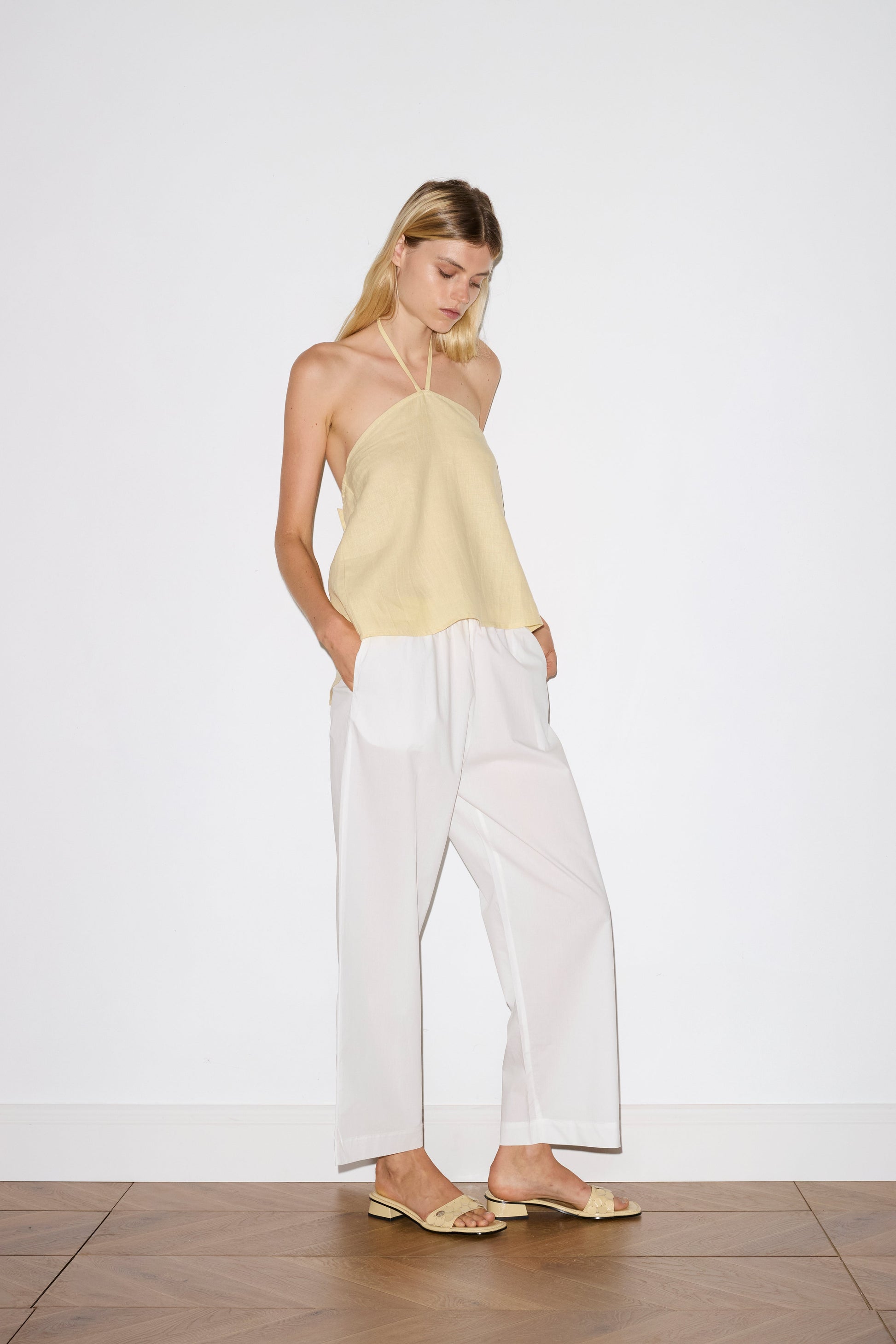 blonde haired female model wears a yellow relaxed halter top features flattering straight neckline, fine tie halter straps and snap closure at back with button detail, worn with a white  ankle length pant features a soft minimal elastic waist with side seam pockets in a straight leg.
