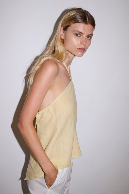 A woman with long blonde hair stands sideways against a white wall, wearing the Linen Wrap Back Top in Butter Yellow and white pants with her hands in her pockets, looking seriously at the camera.