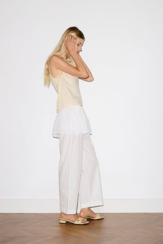 A woman with long blonde hair stands sideways against a white wall, wearing a light beige sleeveless top, the Ease Trouser - White by Deiji Studios, a white layered skirt, and light sandals on a wooden floor. She holds her hands to her head.