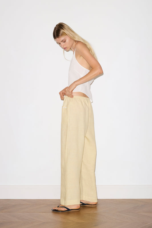 A person with long blond hair, wearing a white sleeveless top, black sandals, and the Linen Square Pocket Pant in light beige, stands indoors on a wooden floor, slightly turned to the side as they adjust their waistband.