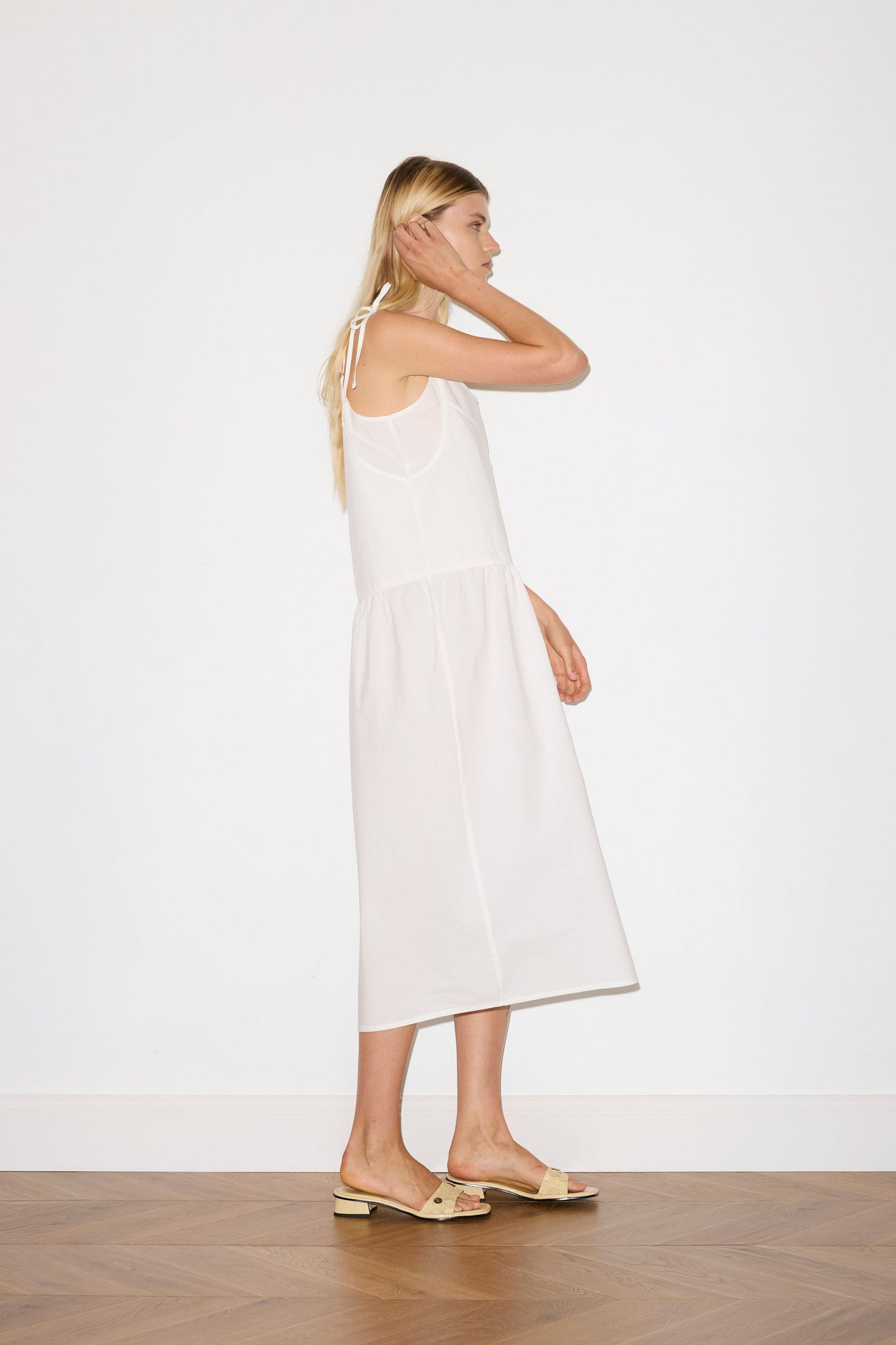 blonde female model wears a white mid-length dress featuring adjustable underlayer straps with sliders for a customised fit and a tied overlayer strap detail at the shoulder