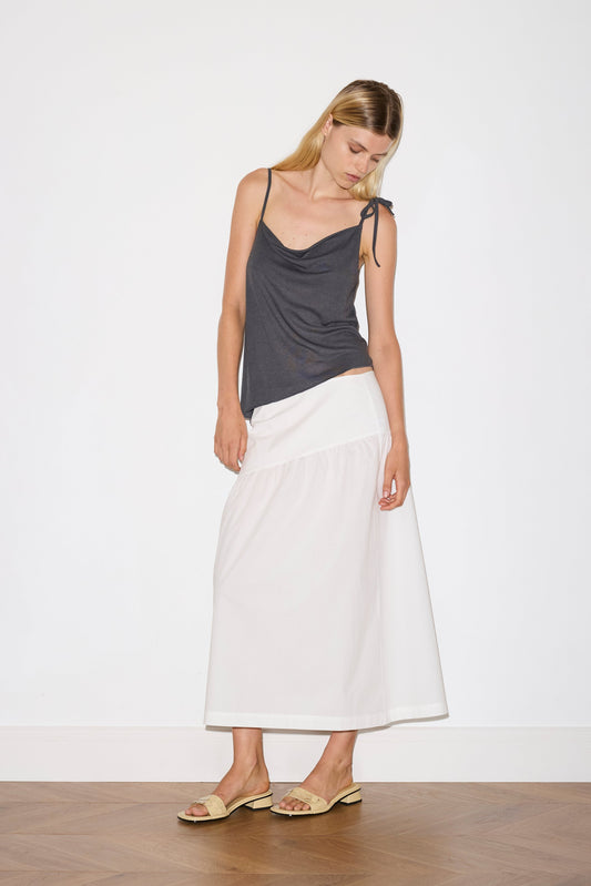 A woman stands indoors against a plain white wall and wooden floor, wearing the Angle Skirt—a long white skirt—paired with a dark gray sleeveless top with thin straps and beige sandals. She looks down, her head gently tilted.