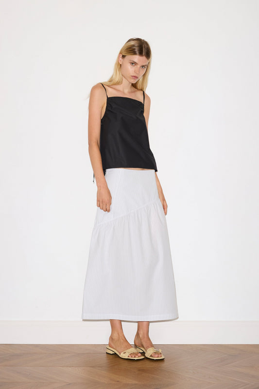 A woman with straight blonde hair and a neutral expression stands indoors against a white wall, wearing a black sleeveless top, the Angle Skirt - Sage Stripe by Deiji Studios, and beige sandals.