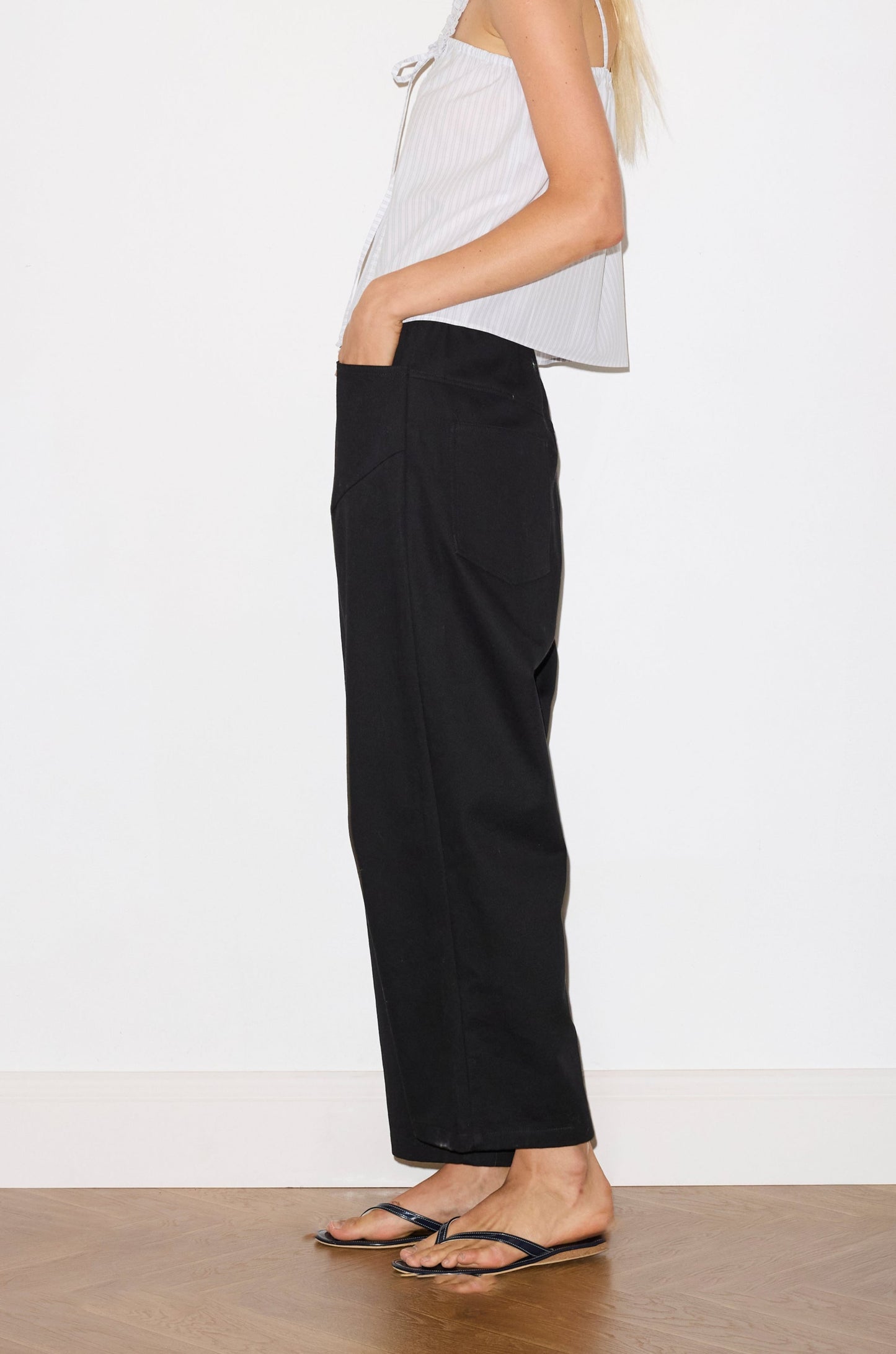 blonde haired female model wears a black Heavy- weighted relaxed-fit pant featuring a wide-leg silhouette, elasticated waistband, and square pocket detail, worn with a sage and white striped a relaxed-fit top featuring self-fabric ruffles along the straps and center front, with a tie front detail. designed with elasticated gathering for ease and comfort