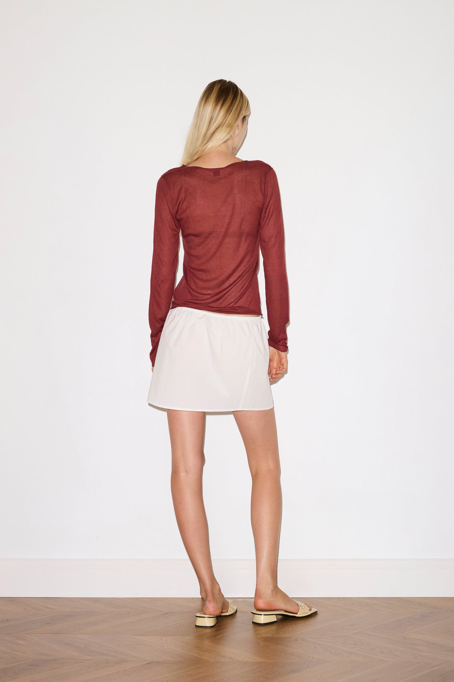A woman with long blonde hair faces a white wall, wearing the 0 Ruffle Mini Skirt - White, a sheer burgundy long-sleeve top, and beige flats on a wooden floor.