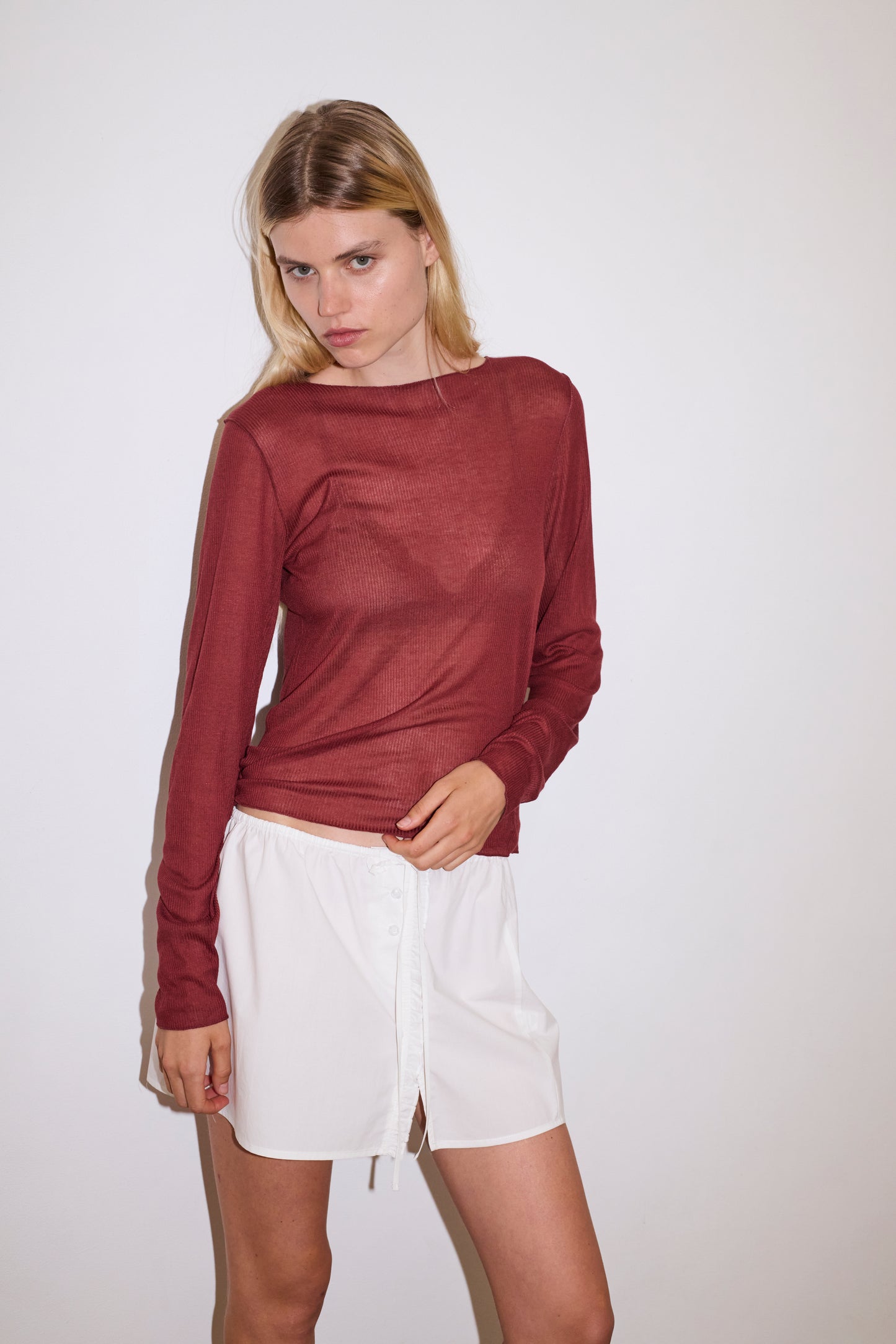 A woman with long blonde hair wears a maroon long-sleeve top paired with the Ruffle Mini Skirt - White by 0, standing against a plain white background.