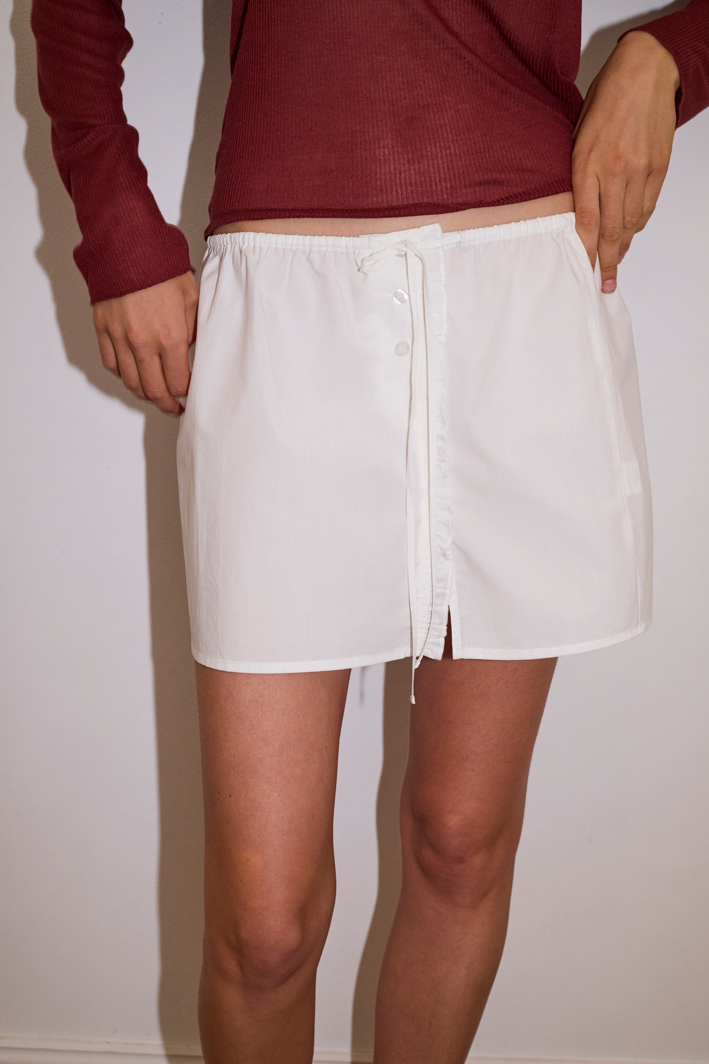 A person wearing the Ruffle Mini Skirt - White by 0, a short organic cotton skirt featuring ruffles, a front tie, and buttons, stands against a plain light wall. Their maroon long-sleeve top is visible but their head is not shown.