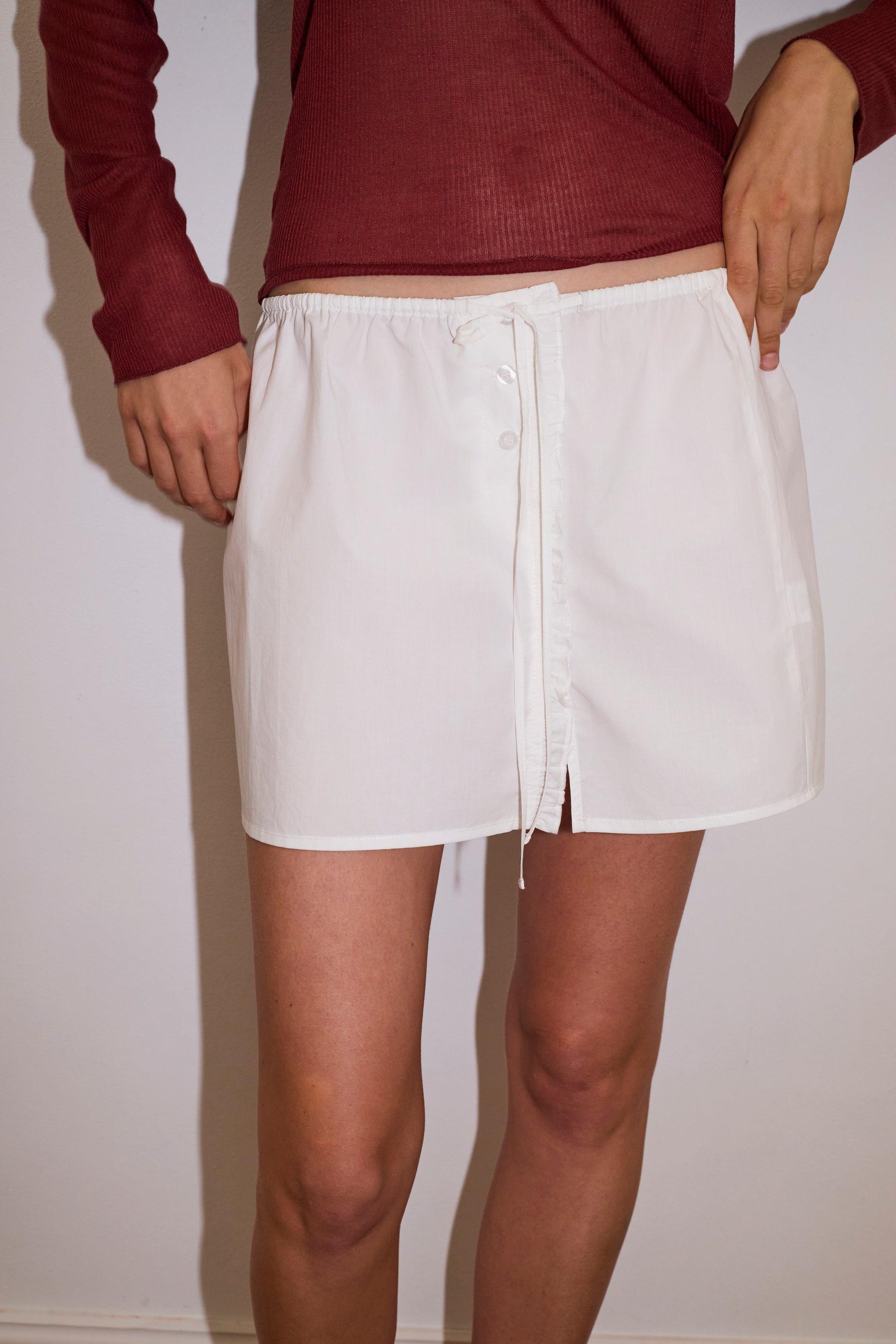 A person wearing the Ruffle Mini Skirt - White by 0, a short organic cotton skirt featuring ruffles, a front tie, and buttons, stands against a plain light wall. Their maroon long-sleeve top is visible but their head is not shown.