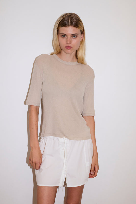 A woman with long blonde hair wears the Split Back Tee - Stone, a beige ribbed short-sleeve top, paired with a white front-button skirt against a plain white background.