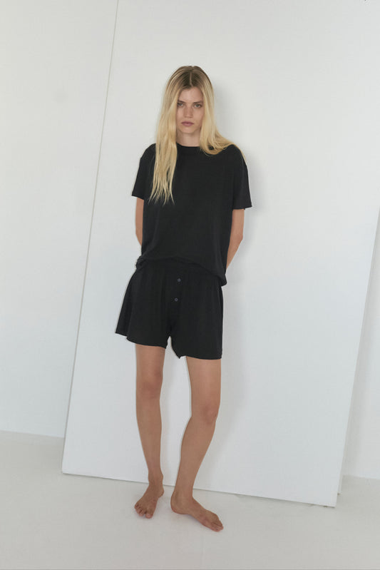 A woman with long blonde hair stands barefoot before a white backdrop, wearing Deiji Studios’ soft t shirt - black and matching shorts, her hands behind her back.