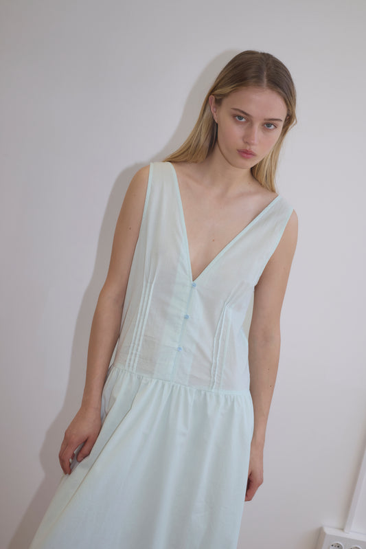 A young woman with long blonde hair wears the Pintuck Dress - Mint by Deiji Studios, a light blue, mid-length dress with a deep V-neck and subtle vertical seams, standing against a plain white wall and looking slightly downward.