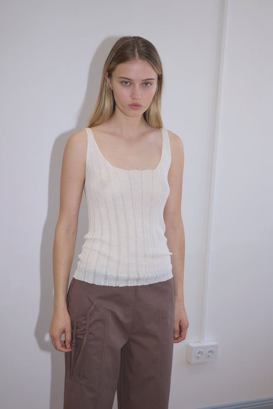 A young woman with straight blonde hair stands against a white wall, wearing the Open Work Tank - White from Deiji Studios and brown zippered pants, looking at the camera with a neutral expression.