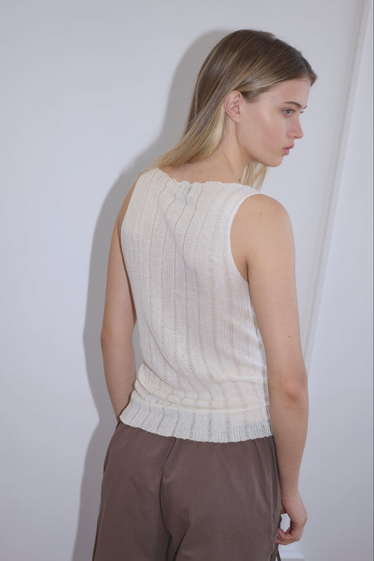 A woman with straight blonde hair stands sideways, looking over her shoulder. She wears the Open Work Tank - White from Deiji Studios and loose brown pants against a plain white background.