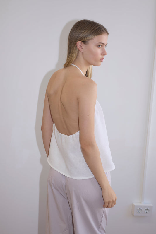 A woman with long, straight blonde hair wears the O Ring Halter - White and light-colored pants, standing sideways against a plain light wall with a visible electrical outlet.