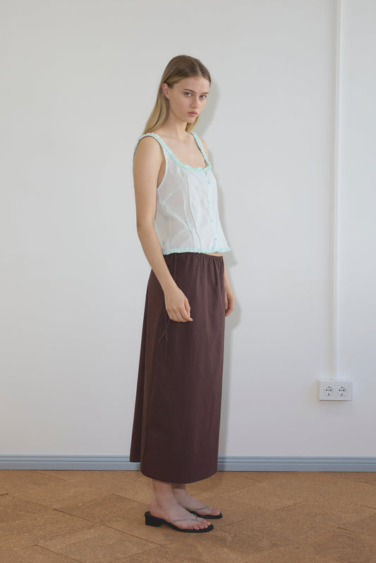 A woman with long blonde hair stands indoors against a white wall, wearing the Button Frill Top - Mint by Deiji Studios, featuring a sheer sleeveless design with ruffled square neckline, paired with a long brown skirt and black flat sandals.