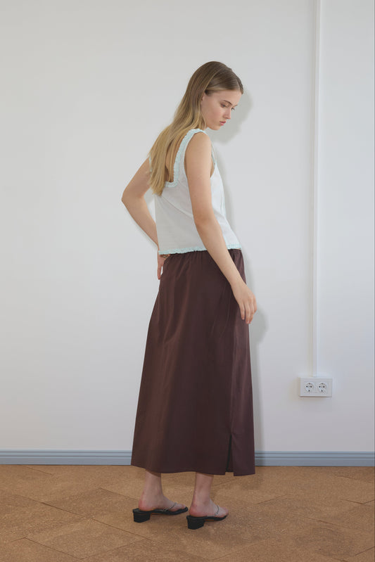 A woman with long blonde hair stands sideways in a minimalist room, wearing Deiji Studios' sleeveless Button Frill Top in Mint, a long brown skirt, and black heeled sandals. She faces left with one hand on her hip.