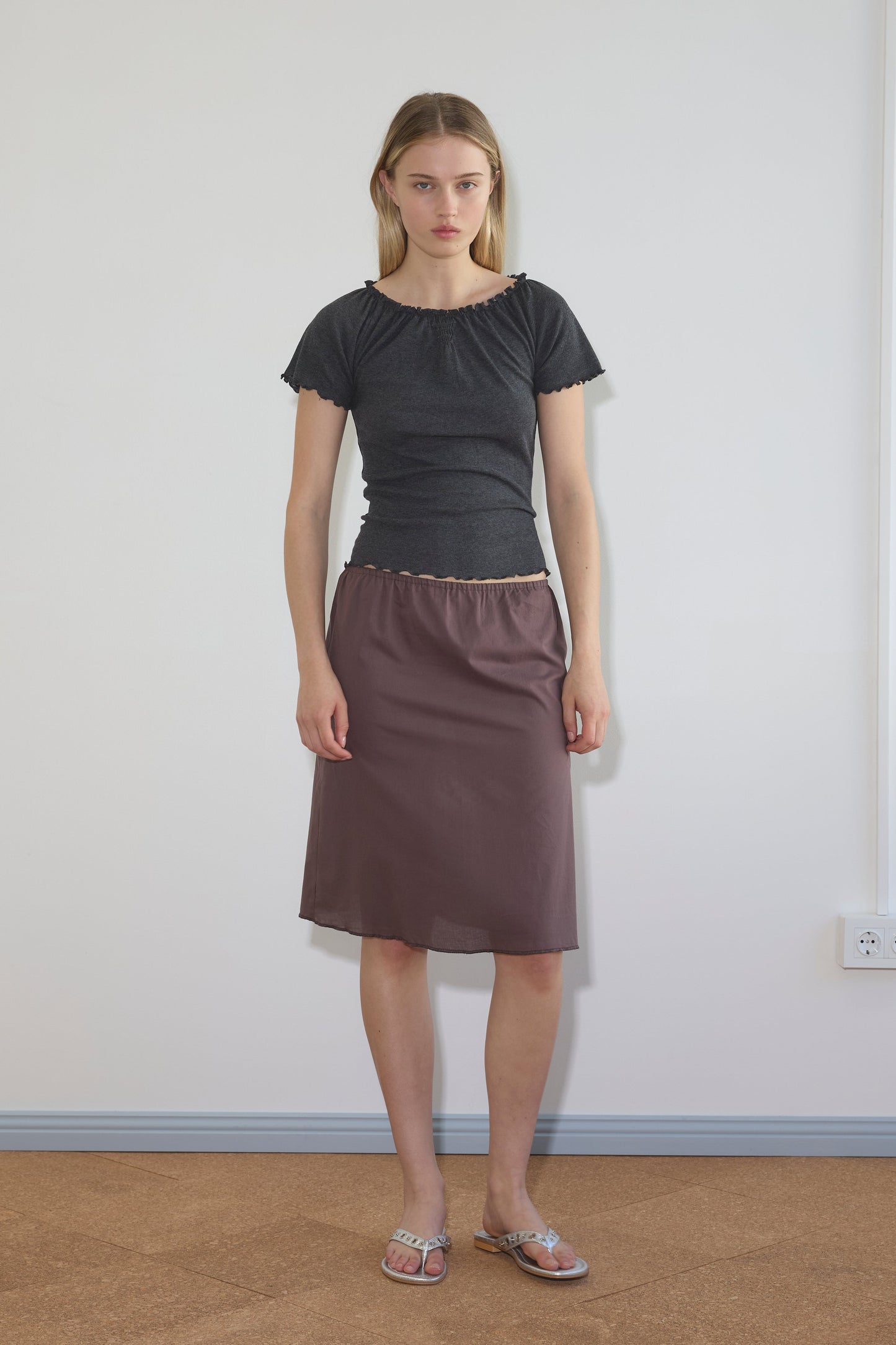 A woman with straight blonde hair and a neutral expression stands indoors against a plain white wall, wearing the Simple Skirt in mauve, a fitted black short-sleeve top, and silver sandals.