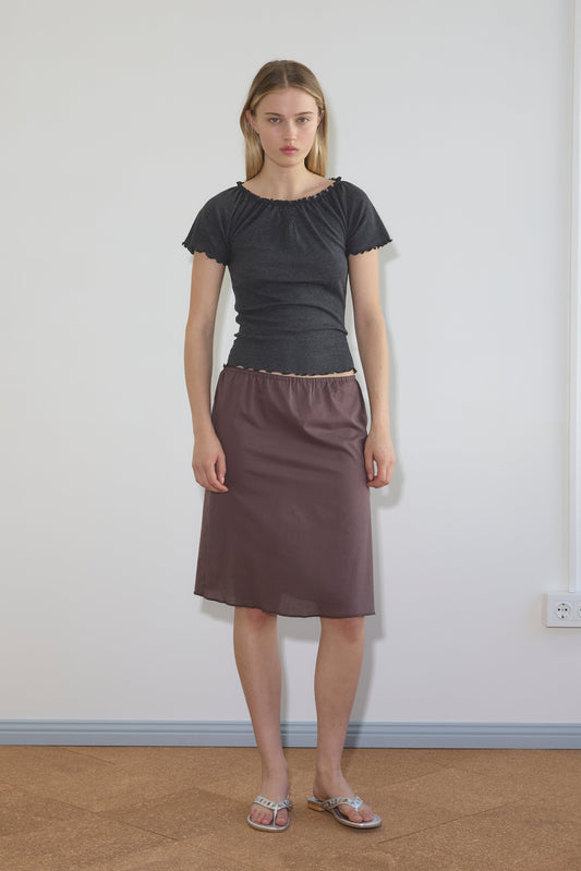 A woman with straight blonde hair and a neutral expression stands indoors against a plain white wall, wearing the Simple Skirt in mauve, a fitted black short-sleeve top, and silver sandals.