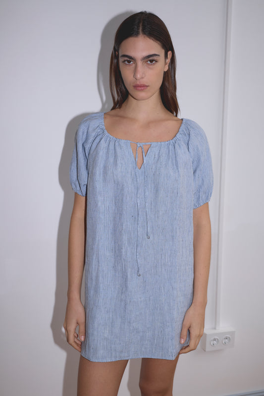 A woman with straight brown hair wears the Smock Dress - Blue Stripe by Deiji Studios, featuring short sleeves and a tie neckline, while standing against a plain white wall.