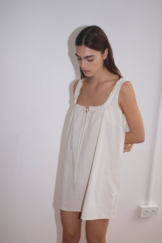 A woman with long dark hair stands against a plain white wall, wearing the Button Frill Mini Dress - Stone Stripe by Deiji Studios. The mini dress features ruffle details and a tie neckline. She looks down with her arms behind her back.