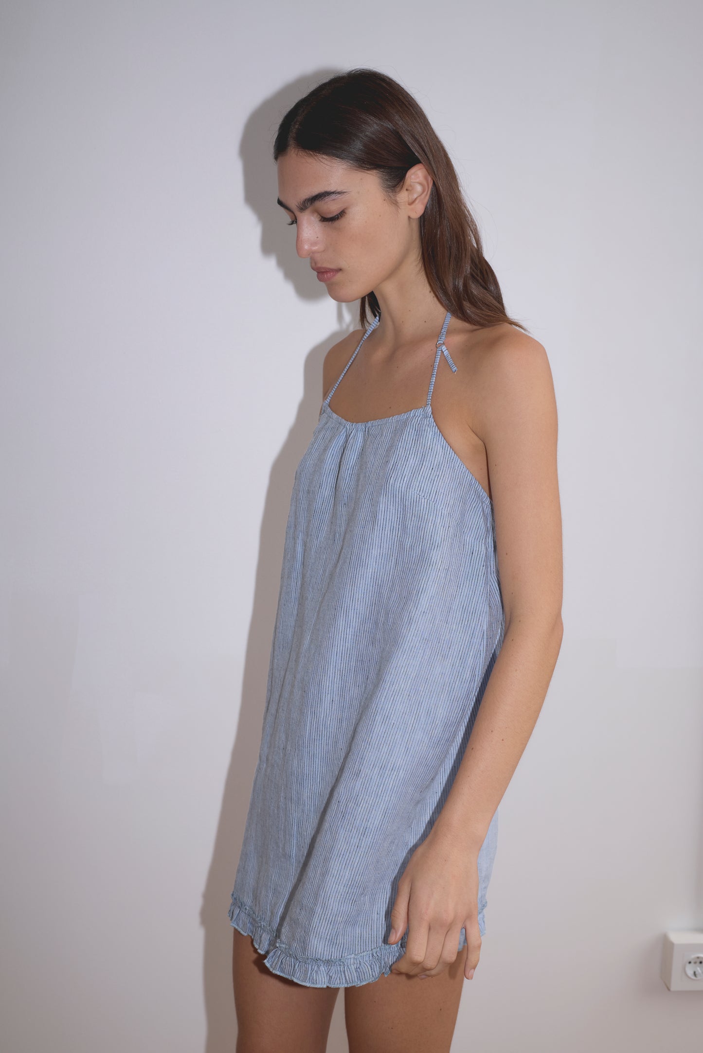 A woman with long brown hair stands against a white wall, wearing Deiji Studios' Flounce Halter Dress - Blue Stripe, a relaxed-fit sleeveless dress featuring subtle vertical stripes and a halter neckline. She looks down with a neutral expression.