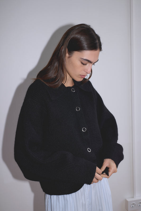 A woman with long dark hair, wearing the Stud Cardi Knit - Black and light blue striped pants, looks down while adjusting her sleeves against a plain white wall.
